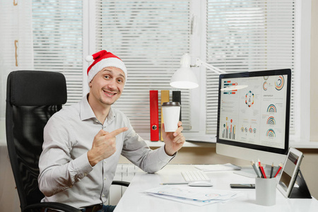英俊的微笑商业男子在衬衫和红色的圣诞帽坐在办公桌上的茶或咖啡, 工作在计算机与现代显示器, 在光办公室的文件, 看着相机
