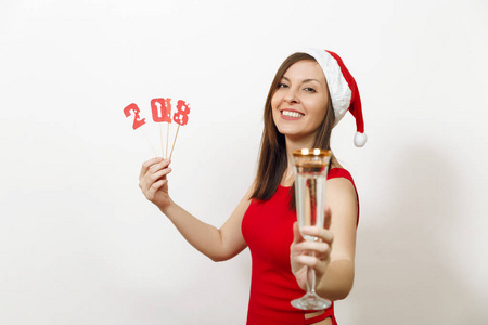 美丽的白种人年轻快乐的女人与迷人的笑容在红色的礼服和圣诞帽持有卡号码2018和在白色背景的香槟杯。孤立的圣诞老人女孩。新年假期