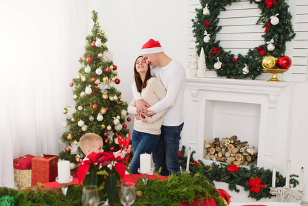 快乐的年轻开朗的父母带着可爱的小儿子戴着红帽子。在家里的小男孩与装饰的新年树和礼品盒的光室。圣诞节好心情。家庭, 爱和假日201