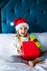 惊喜兴奋的小女孩在圣诞老人的帽子打开目前, 而坐在床上的交叉腿圣诞节早晨