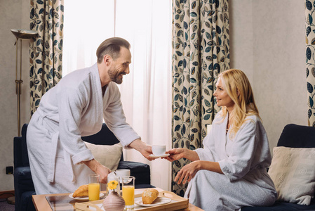 在酒店房间一起吃早餐的同时，在浴袍中相互微笑的快乐成熟的一对