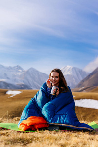 快乐的年轻女孩旅行者坐在睡袋里喝着来自高加索山脉背景的热杯茶