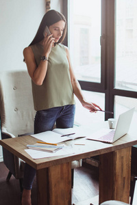 我们需要在这份报告中做一些调整 一位年轻漂亮的女商人站在办公桌前，一边用手机说话，一边看着她的笔记本电脑