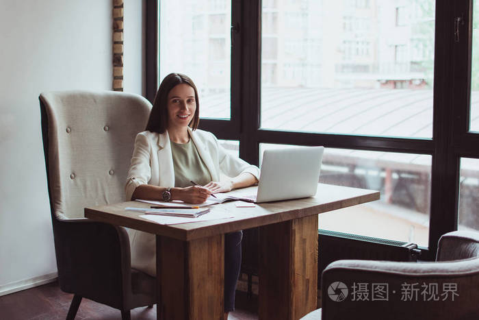 美丽的年轻女商人微笑着写着东西,坐在办公桌前看着相机照片-正版