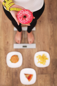年轻漂亮的女人站在磅秤上, 拿着一个甜甜圈。健康饮食的概念。健康的生活方式。饮食
