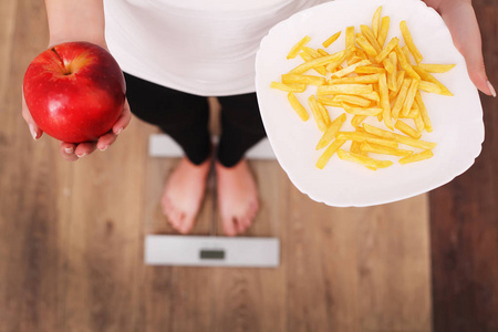 一个年轻漂亮的女人在磅秤上, 在苹果和薯片之间做出选择。健康饮食的概念。健康的生活方式。饮食