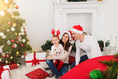 快乐开朗的父母带着可爱的小儿子。小男孩戴着红帽子, 坐在家里的灯房里, 带着装饰一新的圣诞树和礼品盒。圣诞节好心情。家庭, 爱和