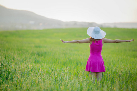 年轻女子穿着紫色的衣服，戴着白色的帽子，走在绿色的田野上，背景是山。