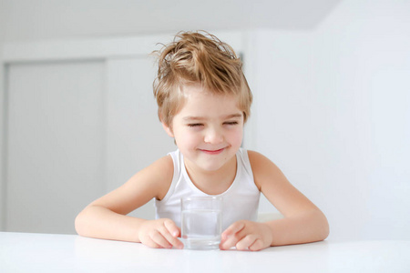 有趣的小男孩坐在白桌旁，端着一杯水