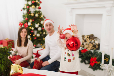快乐的年轻父母带着可爱的小儿子在帽子里。小男孩带着大红树的玩具球站在装修的新年灯房里。圣诞节好心情。家庭, 爱和假日2018概念