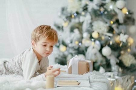 小男孩躺在圣诞树旁的白色床上，在笔记本上写字