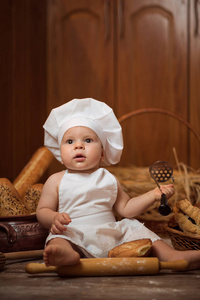快乐的小孩穿着厨师服装在厨房里坐在桌子上，篮子里放着美味的面包卷和面包。 复制空间