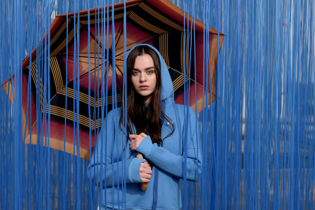 年轻漂亮的女人带着橙色的伞，时髦地摆在蓝色的背景上。 雨的概念