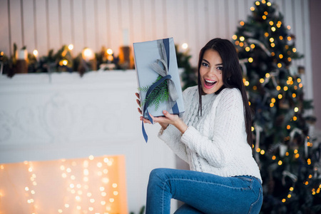 穿着毛衣的年轻微笑的妇女拿着礼物盒庆祝冬天假日在装饰的家庭内部与圣诞树。新年快乐理念