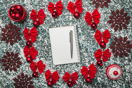 从上面的记事本和笔在一个圆圈内，由红色蝴蝶结常绿玩具与冬季雕像在灰色大理石背景上创建，有空间写文字问候。