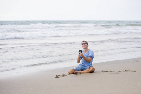 迷人和英俊的男子在他的30s 坐在沙滩上放松在海滩上笑着在海边发短信的手机