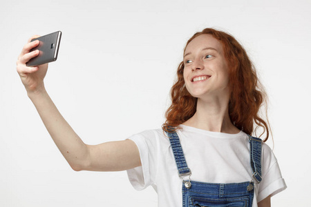 照片的美丽年轻红发女性孤立的灰色背景, 伸展手臂与智能手机采取自拍图片分享它在社会网络