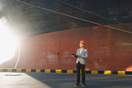 英俊的胡子拉碴的成功商人穿着灰色西装, 防护建筑橙色头盔的手持碑, 站在海港对货物生锈的船的背景。男性与小工具在晴朗的天