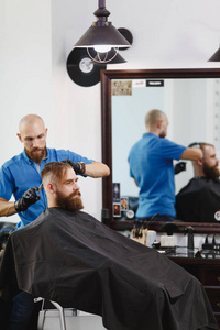 男专业理发师用剪刀为客户服务。姜英俊的野蛮时髦的年轻人与厚实的大胡子, 短的头发在黑海角得到时髦发型。轻的白色理发店房间
