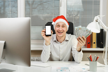 微笑的愉快的商人在衬衣, 红色圣诞节帽子坐在书桌与移动电话和许多现金金钱, 工作在新的年在计算机与现代显示器, 文件在轻的办公室