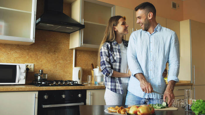 快乐对年轻情侣接吻拥抱和聊天在厨房里做饭早餐在家时