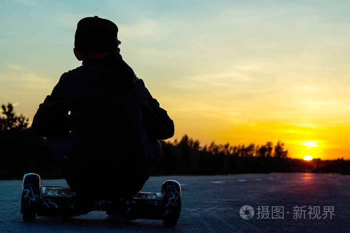 一个小男孩坐在一辆超级流行的电动摩托车上, 看着日落。迷你赛格威气垫船运输装置, 广受欢迎的城市交通