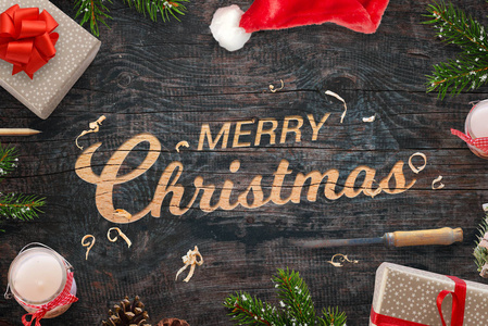圣诞快乐的问候刻在木头表面。 圣诞礼物树枝凿蜡烛针和圣诞老人的帽子旁边。