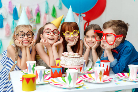 滑稽的孩子们戴着派对帽, 大眼镜, 在生日蛋糕上看着大胃口, 想尝尝它
