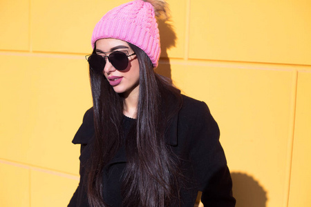 美丽的黑发女人戴着太阳镜和针织帽。时尚的年轻博客女性，蓝色头发，穿着休闲风格的衣服，黑色夹克，粉红色的帽子，太阳镜摆在黄色的墙上