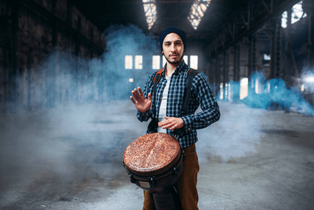 年轻的男性鼓手演奏非洲木制鼓烟和工厂的背景。 Djembe音乐打击乐器