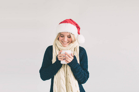 戴着一杯咖啡的圣诞老人微笑的女人, 在白色的隔离。女孩穿着毛衣, 圣诞帽和围巾工作室拍摄