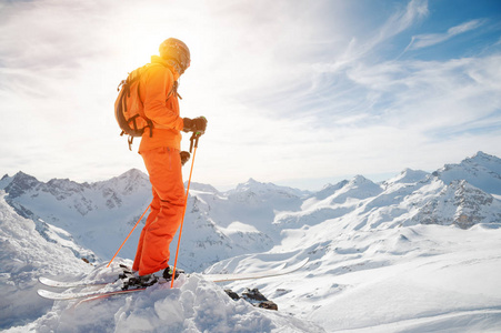 在一个美丽的背景下, 在一个白雪皑皑的深渊前的悬崖上, 一个在他的手上背着一个背包, 戴着头盔和滑雪杖的橙色的滑雪者。