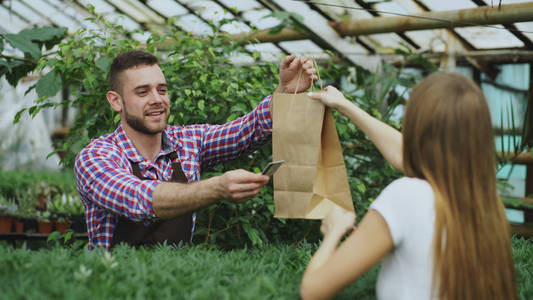 年轻微笑卖方花店在花园的中心工作。男人给客户和付款用信用卡在商店提供的购物袋
