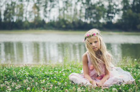 美丽的白种人女孩，一头金色的长发，穿着粉红色的连衣裙，独自坐在河边和树林旁的绿草中，带着悲伤的表情