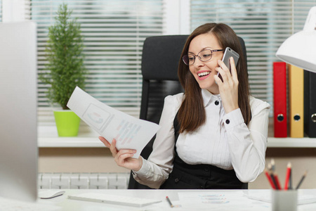 美丽微笑的商业妇女在西装和眼镜坐在办公桌上, 在现代电脑工作, 在轻型办公室的文件, 在手机上交谈, 进行愉快的谈话