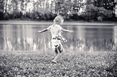 一个金发碧眼的小女孩，穿着白色连衣裙，在河边的绿草中跳舞，单色