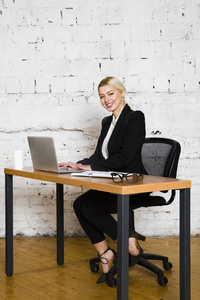 年轻的金发美女女实业家坐在办公室的桌子上, 笔记本电脑, 笔记本和眼镜的西装。经营理念