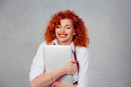 我喜欢我的笔记本电脑。红头卷发商业妇女在白衬衫拥抱电脑微笑快乐的眼睛闭上灰色背景墙