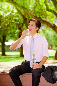 英俊的年轻商人穿着紫色的 tshirt, 吃着三明治, 用他的另一只手在户外拿着一瓶水, 在一个模糊的公园背景