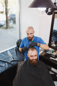 男专业理发师用剪刀为客户服务。姜英俊的野蛮时髦的年轻人与厚实的大胡子, 短的头发在黑海角得到时髦发型。轻的白色理发店房间