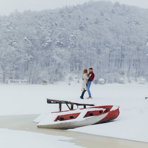 Lovinghappy 夫妇在靠近小船和森林的雪地草地上拥抱和玩乐。冬季时间