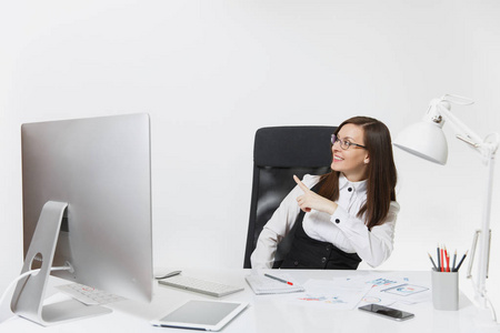 微笑的商业妇女坐在办公桌前, 工作在计算机与文件在光办公室, 指着她的手指在白色背景与复制空间为您的广告或促销内容