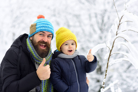 大拇指从父亲和儿子。大胡子男子和开朗快乐的男孩一起微笑和显示手指在冬季公园附近的滑雪胜地的背景。彩色帽子的滑雪者。冬季运动
