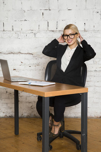 年轻的金发美女女实业家坐在办公室的桌子上, 笔记本电脑, 笔记本和眼镜的西装。经营理念