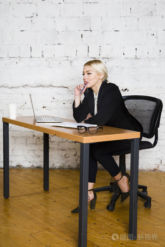 年轻的金发美女女实业家坐在办公室的桌子上, 笔记本电脑, 笔记本和