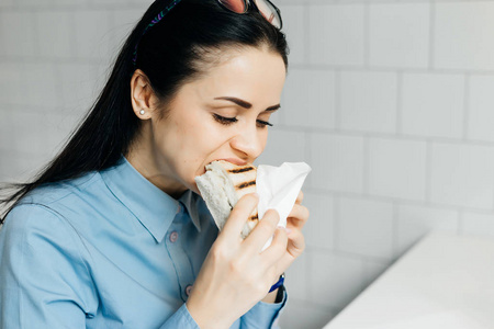 饥饿的年轻黑发女孩在蓝色衬衣急切地吃三明治在咖啡馆