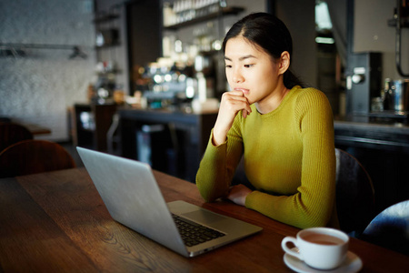 年轻的忧郁的女性坐在笔记本电脑前的咖啡馆里，阅读在线信息
