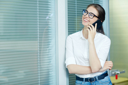 穿休闲服和戴眼镜的年轻女性，在办公室用智能手机说话