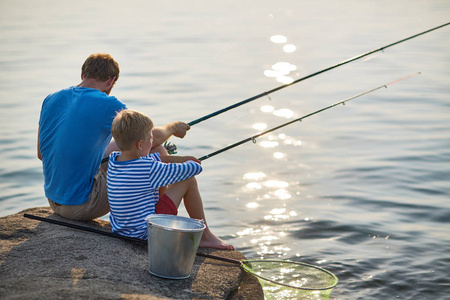 在阳光明媚的夏日里，一个成年人和一个十几岁的男孩坐在一起，在平静的蓝湖波光粼粼的水域里钓鱼