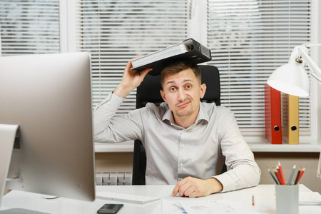 震惊困惑和压力的商人坐在办公桌前的衬衫, 在电脑上工作与现代显示器, 文件夹, 灯, 文件在轻办公室的窗口背景。经理或工作人员